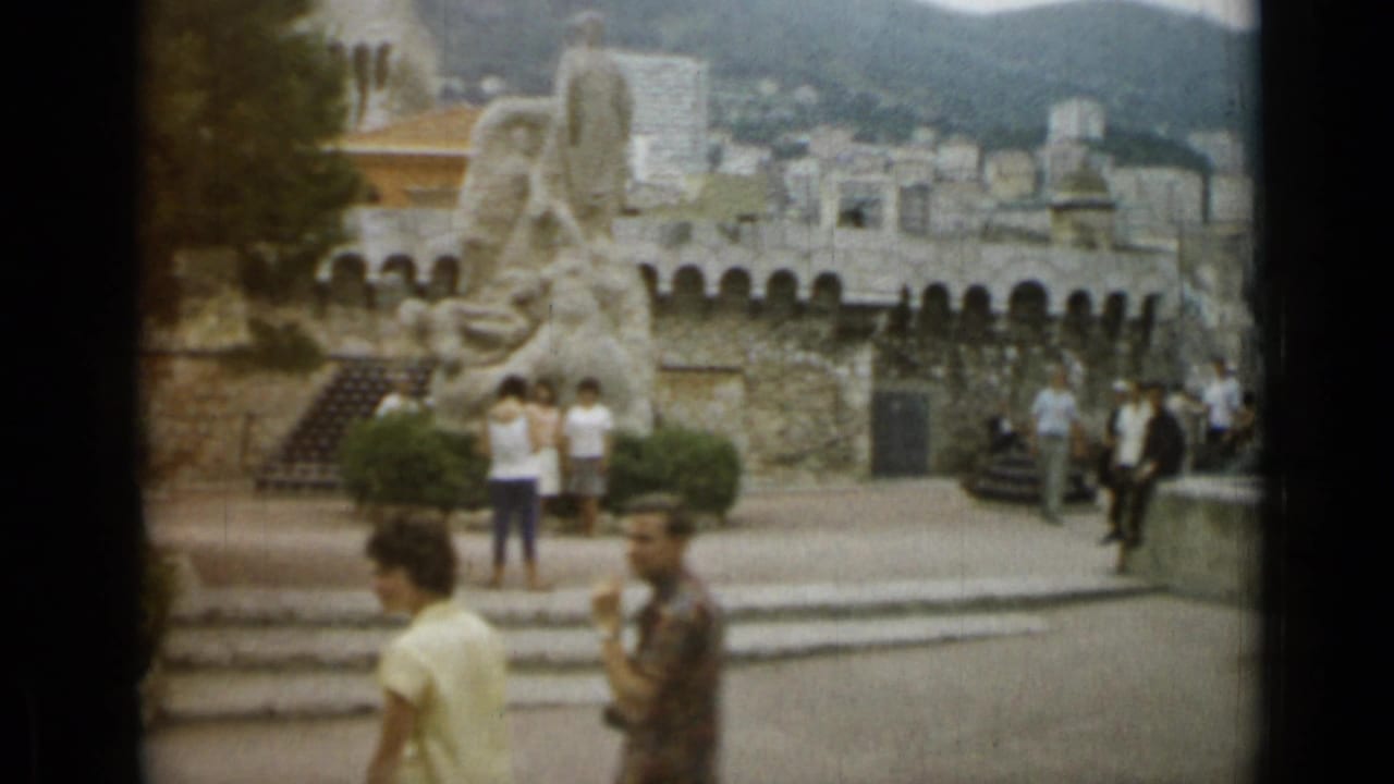 Tourists in summer clothing pass through a Nice public square near a large stone monument, steps, and distant hillside buildings.