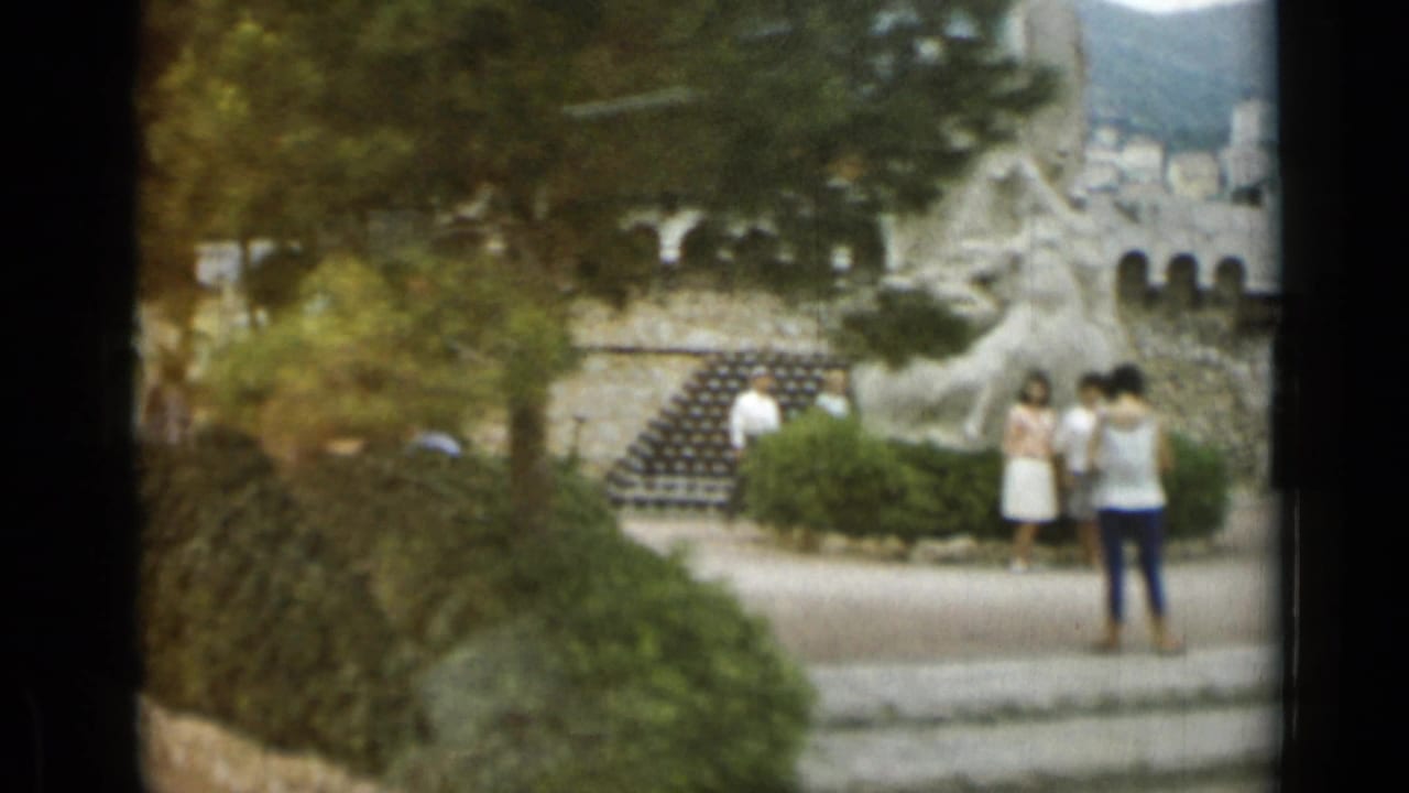 Nice, France 8mm frame with visitors pausing near landscaped shrubs, a pale stone monument, steps, and an old wall.