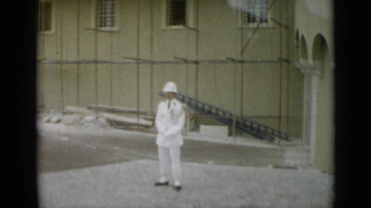 White-uniformed guard standing in a paved courtyard beside pale stone buildings.