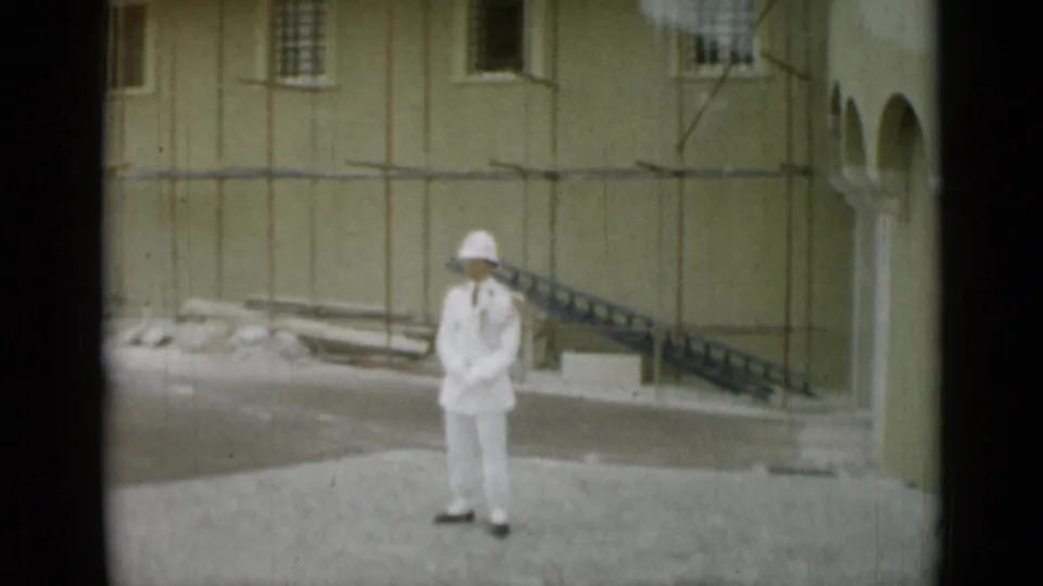A white-uniformed guard stands in a courtyard at a guarded site in Nice, France, in 1962, with pale walls and barred windows behind him.