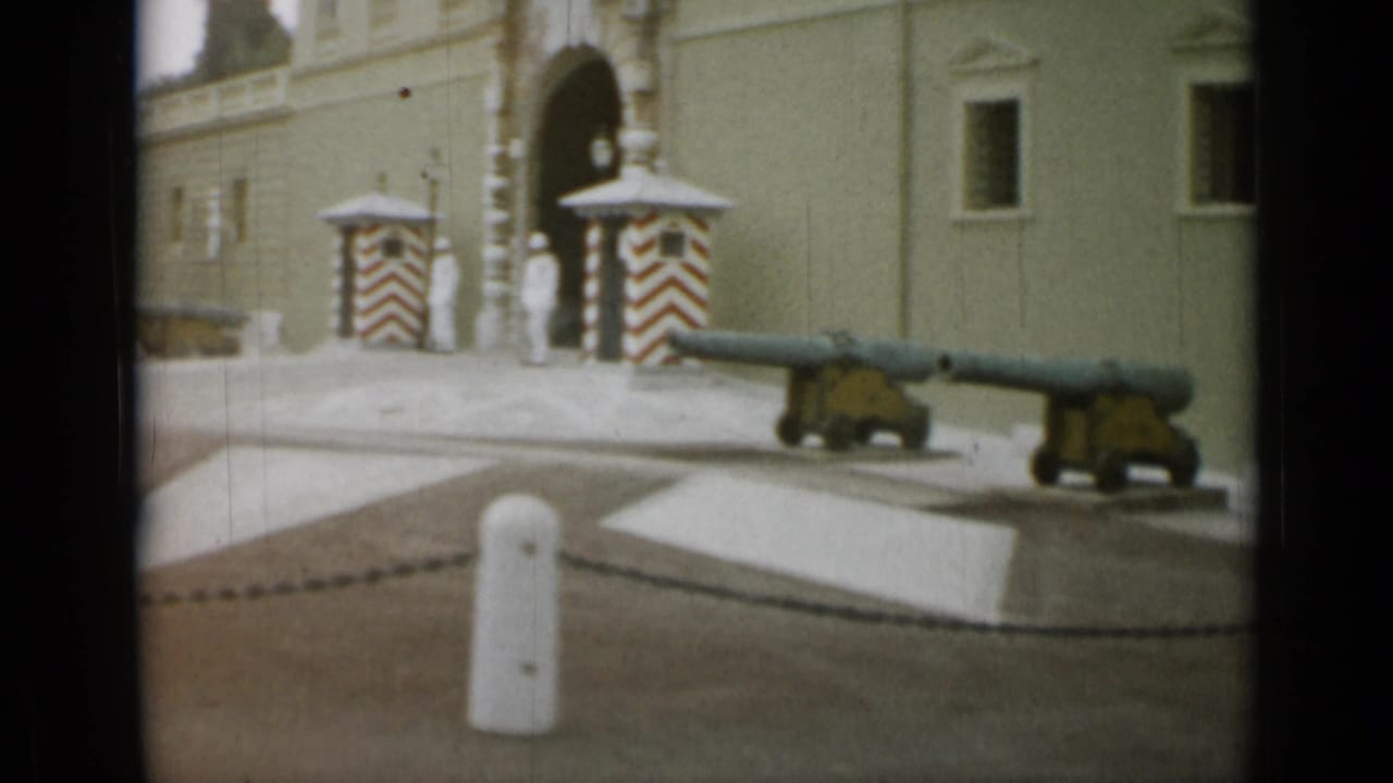 A guarded arched entrance with striped sentry boxes, white-uniformed figures, cannons, bollards, and chain barriers is seen in the Nice, France clip.