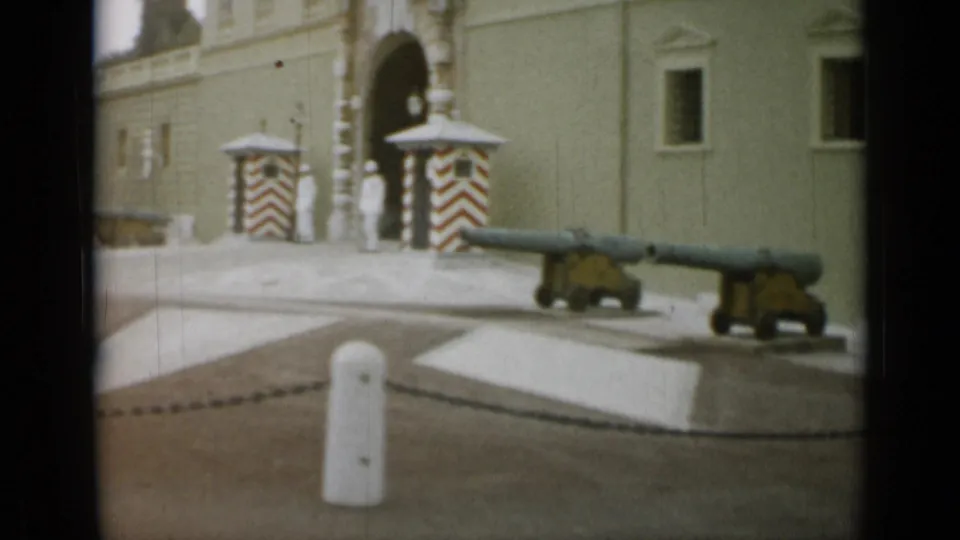 A guarded arched entrance with striped sentry boxes, white-uniformed figures, cannons, bollards, and chain barriers is seen in the Nice, France clip.