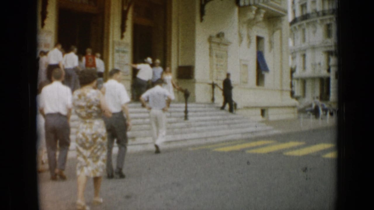 Tourists and pedestrians gather near the steps of a prominent building in 1962 Nice, with street markings visible in the foreground.