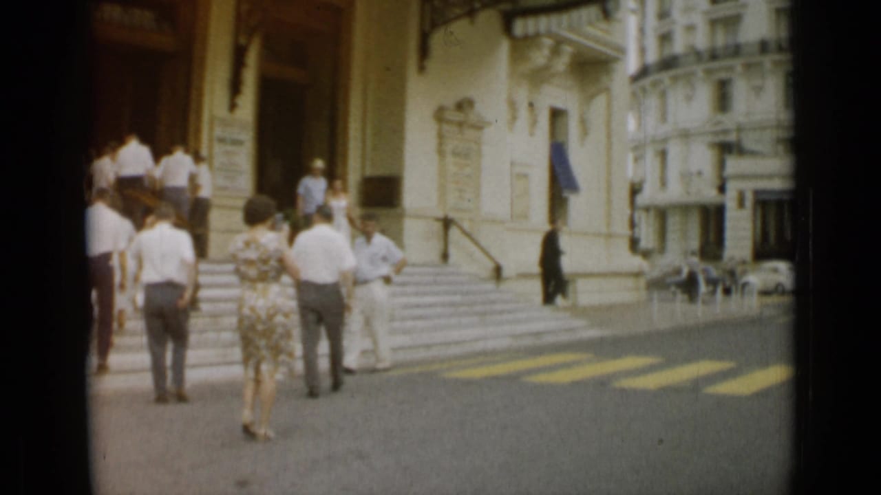 Tourists remain near the entrance steps in 1962 Nice as pedestrians pass along the sunlit street and crosswalk.