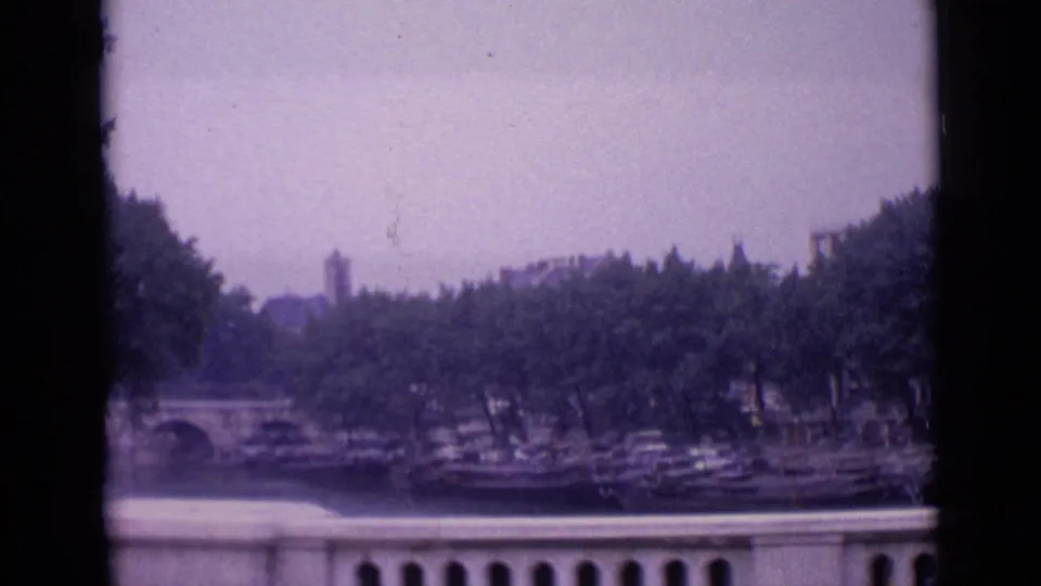 1962 Paris home-movie frame showing a hazy waterfront view with bridge arches, docked boats, trees, and a stone balustrade.