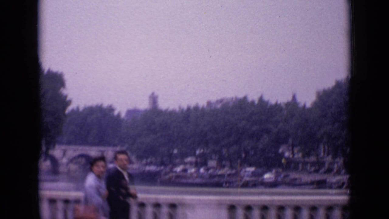 1962 Paris home-movie frame with two pedestrians by a balustrade overlooking a waterway, bridge, boats, trees, and parked cars.