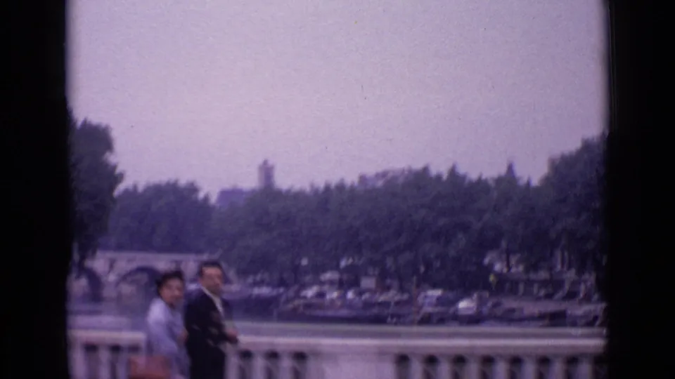 1962 Paris home-movie frame with two pedestrians by a balustrade overlooking a waterway, bridge, boats, trees, and parked cars.