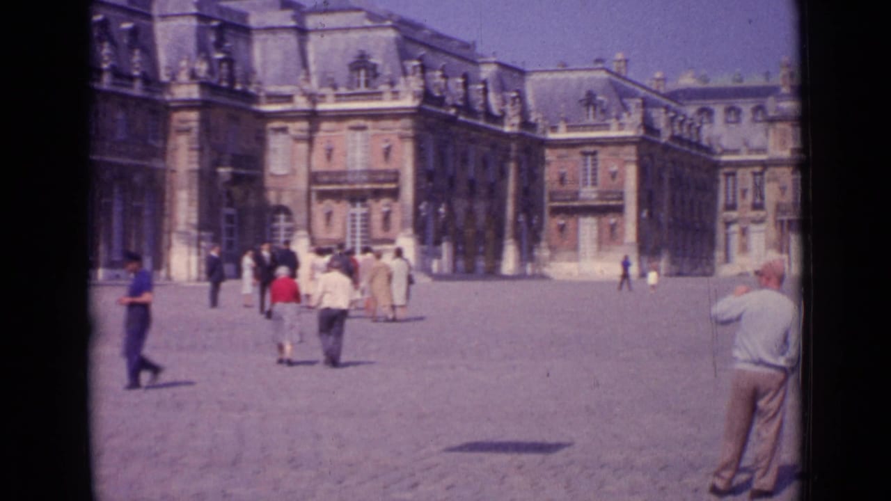 Tourists cross a broad paved plaza in front of ornate multi-storey buildings.