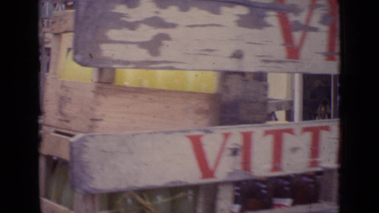 Close 1962 Paris home-movie frame of stacked crates with partial Vittel branding, bottles, and shopfront details in soft focus.