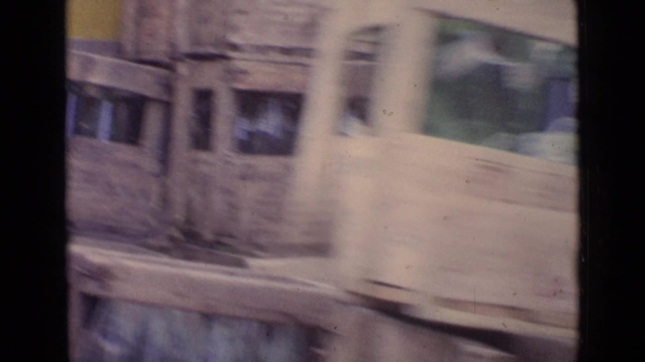 Motion-blurred 1962 Paris street view with a passing light-colored vehicle, storefront glass, and building facade details.
