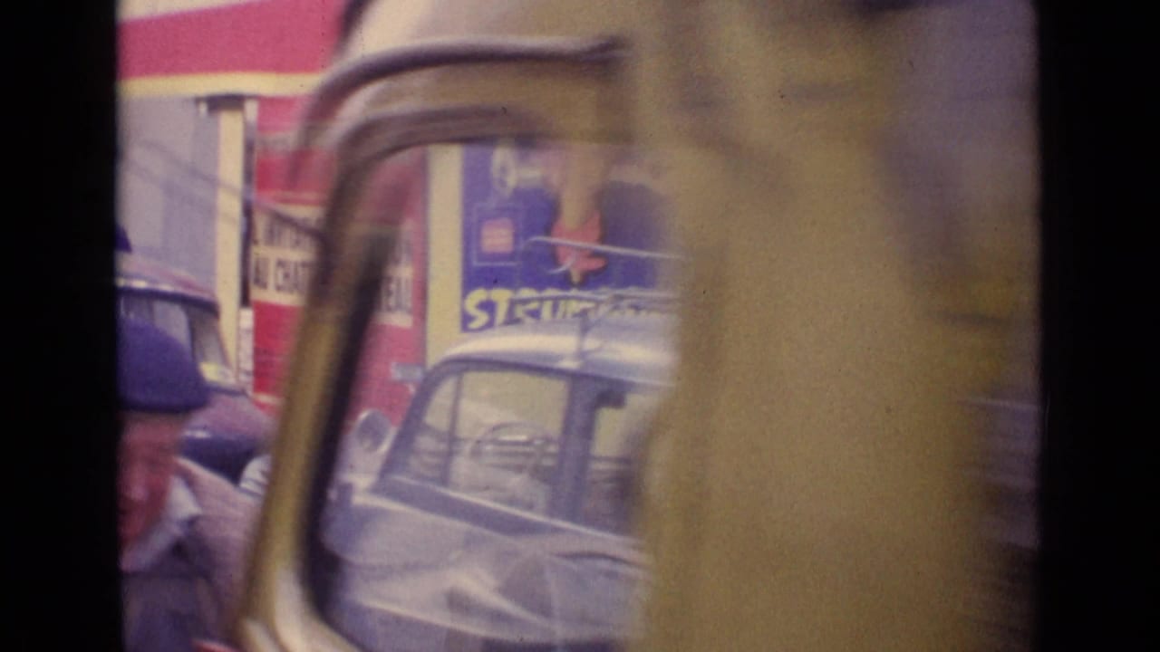 Paris street seen past a vehicle window with vintage cars, a pedestrian in a beret, and a storefront sign.