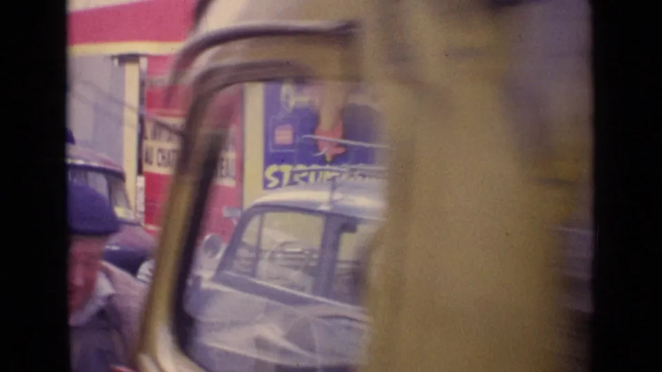 1962 Paris street view with vintage cars, a man in a beret, storefront advertising, and partial vehicle framing in the foreground.