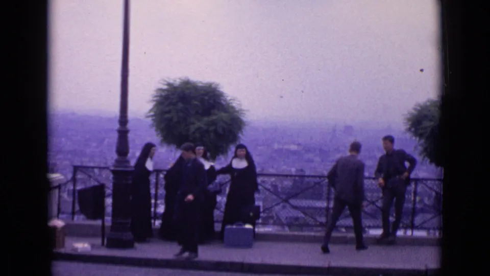 1962 Paris 8mm frame of nuns and male visitors near a scenic railing, framed by a lamppost and tree planter above the city.