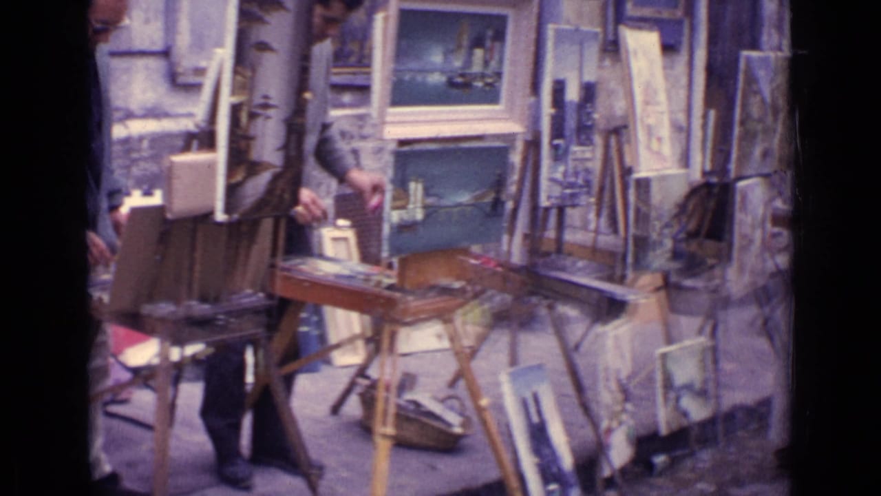 Outdoor art display with easels, framed paintings, and a painter partly visible on a cobblestone street.