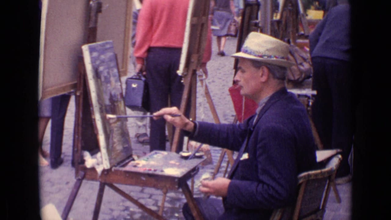 Man in a straw hat paints at an easel beside a palette and framed canvases on the street.