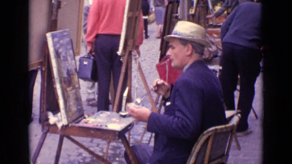 Adult street painter in a hat sits at an easel in Paris, holding a palette and brush while pedestrians pass behind him.