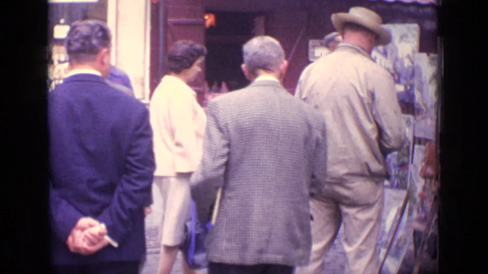 Pedestrians in 1962 Paris gather near a street display of artwork, including a tall man in a hat and others facing the stall.
