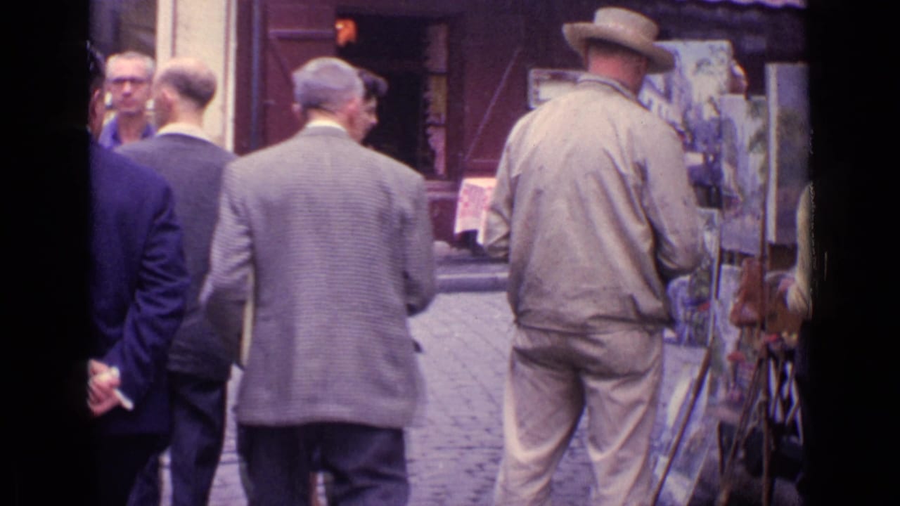 Street painter seen from behind as suited pedestrians pass near outdoor easels and a cafe frontage.