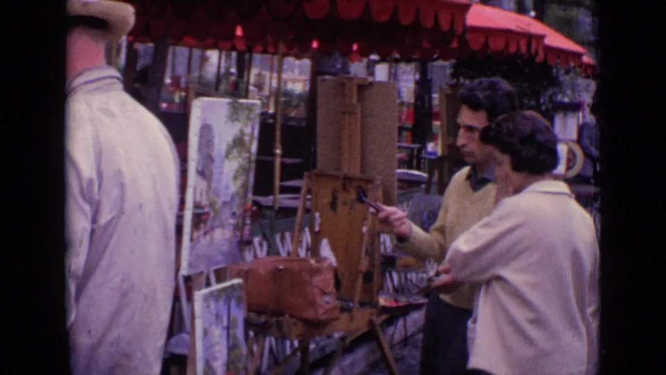 1962 Paris street-art setup with a man and woman beside easels, brushes, a wooden paint box, and paintings beneath a red awning.