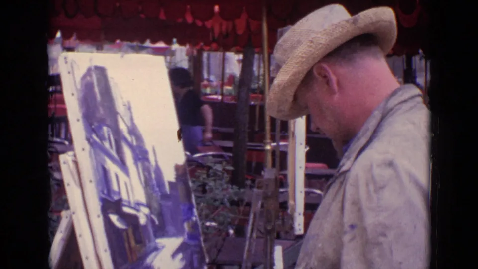A close home-movie view captures the Paris painter leaning toward his cityscape canvas in front of an outdoor café.
