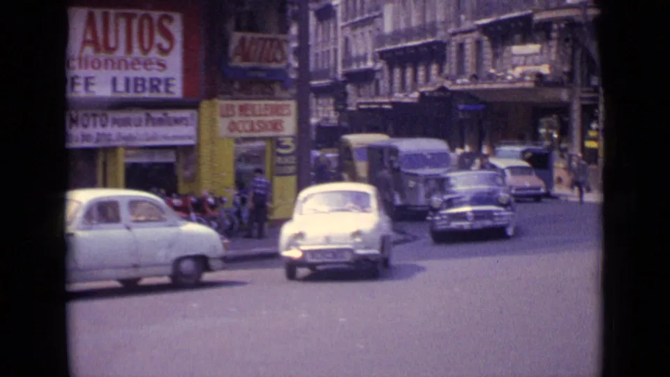 1962 Paris street scene with mid-century cars, storefront signage, scooters, pedestrians, and multi-story buildings.