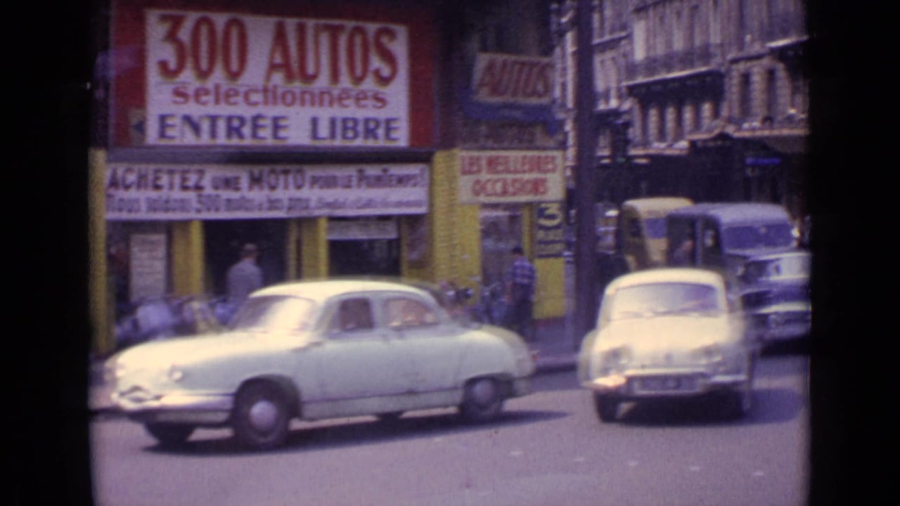 1962 Paris street view with a pale sedan, pedestrians, and large French signs advertising cars and motorcycles.