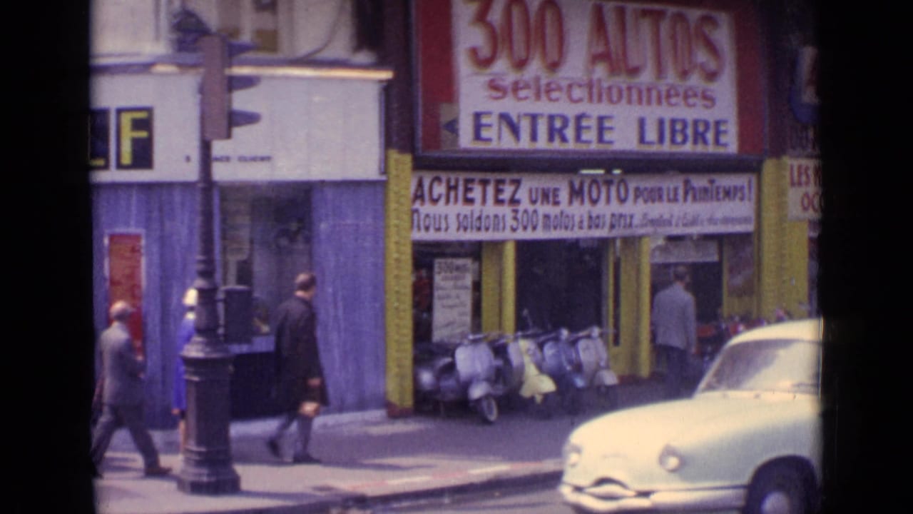 Pedestrians walk past scooters and French auto signage near a street corner.