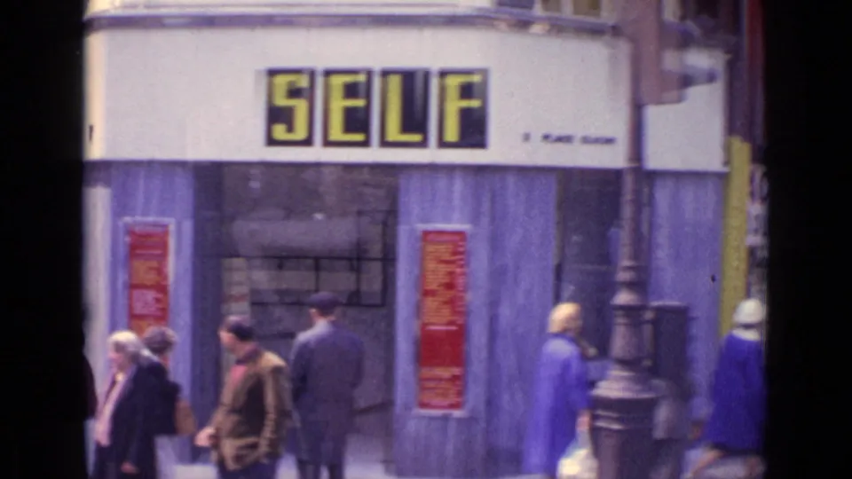 1962 Paris sidewalk scene with pedestrians passing a storefront marked SELF, red posters, and a streetlamp.