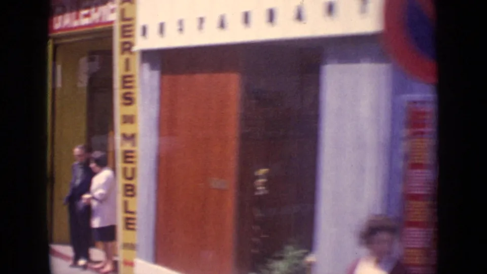 Paris storefront entrance with pedestrians nearby, a vertical furniture sign, and blurred passing street movement.