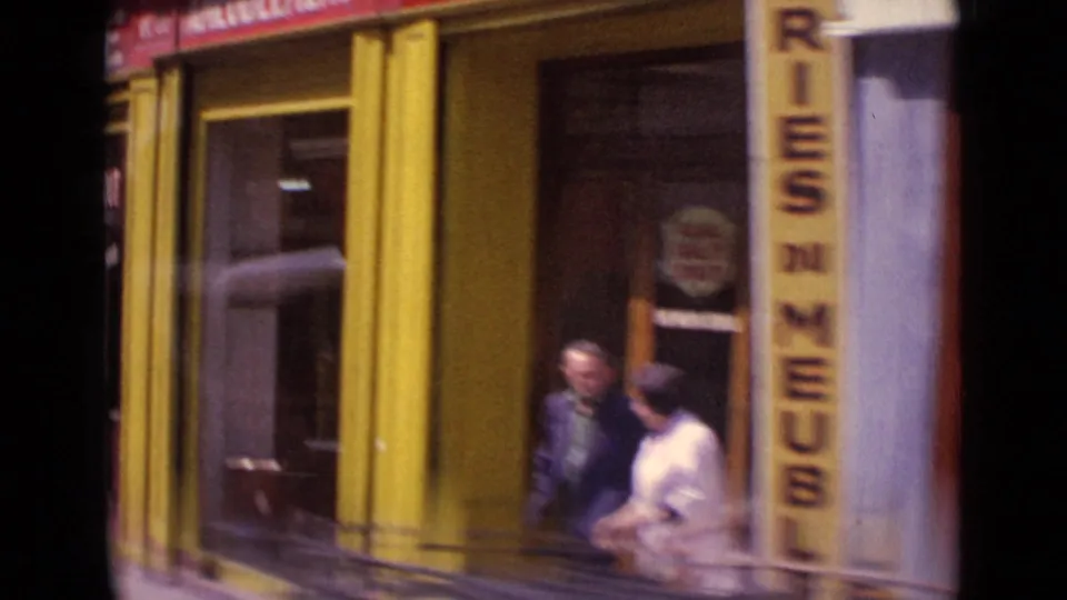 1962 Paris street view with a yellow shopfront, vertical sign, and adults passing near the entrance in soft 8mm color.