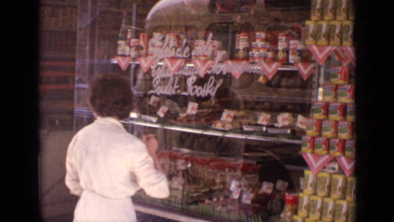 In 1962 Paris, a woman in a white dress looks into a grocery shop window with handwritten signage and stacked food tins.