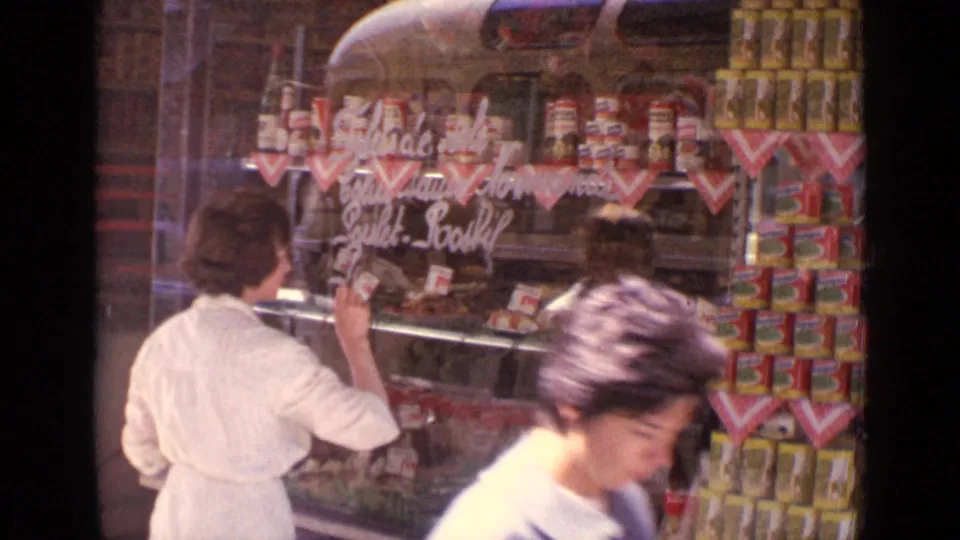 A Paris grocery window display is seen as the woman in white looks on and a passerby moves across the foreground.