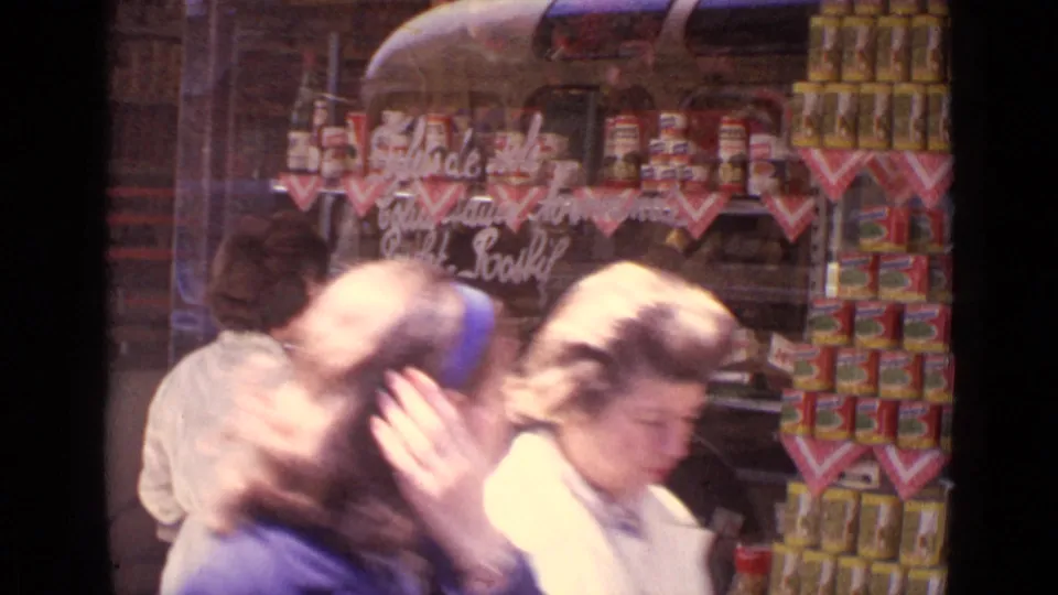 Adults move through the foreground of a 1962 Paris shop-window scene, partially obscuring the woman at the food display.