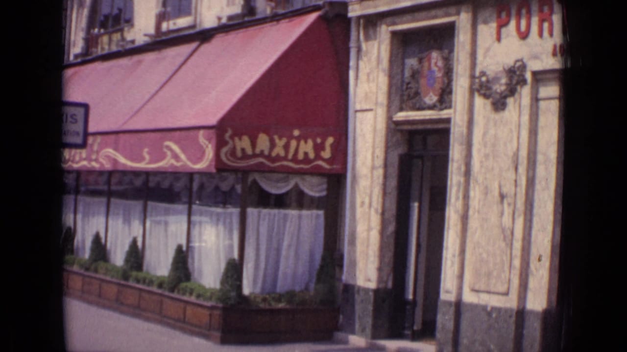 Paris street-level view of Maxim's restaurant frontage with red awning, decorative facade details, curtains, and planters.