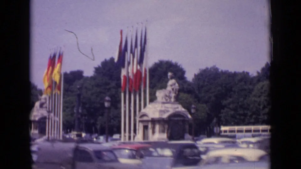 1962 Paris public square view with a flag-lined monument, passing cars, trees, and a bus seen through soft 8mm color.