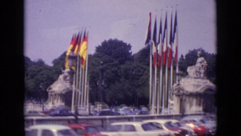 Paris 8mm frame showing a stone monument flanked by national flags, with 1960s cars, trees, and a bus in the surrounding square.