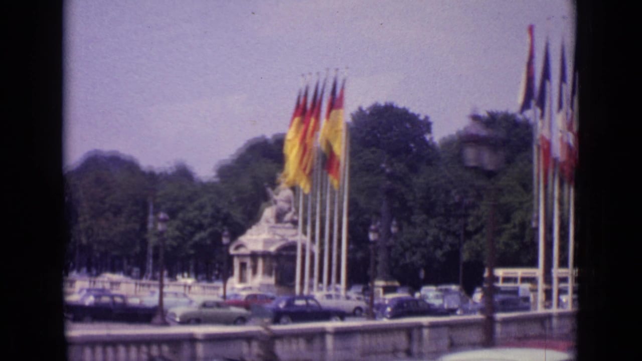 German and French flags on tall poles beside a Paris monument and 1960s cars.