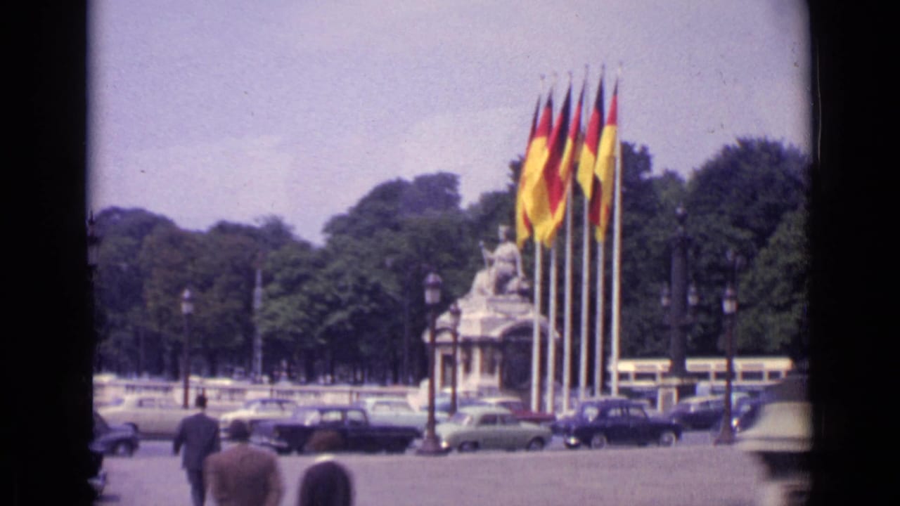 1962 Paris view showing German and French flags near a monument, with pedestrians in the foreground and cars moving through the square.