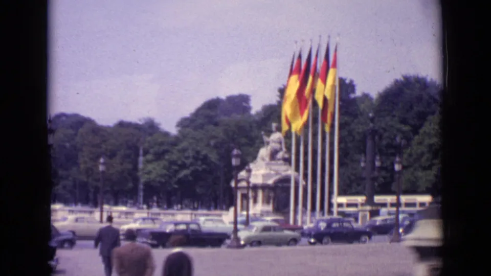 1962 Paris view showing German and French flags near a monument, with pedestrians in the foreground and cars moving through the square.