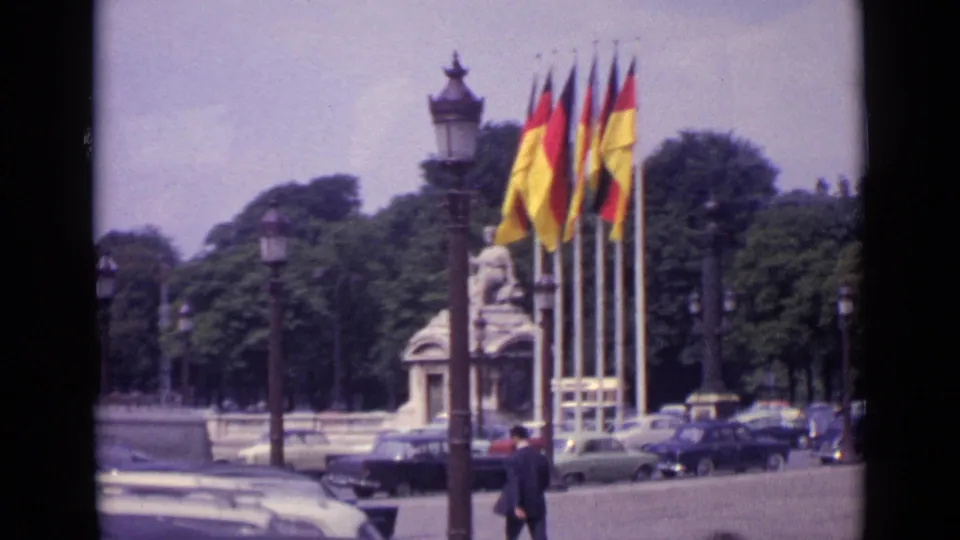 Closer 1962 Paris frame with German and French flags, an ornate lamppost, a stone monument, trees, and parked mid-century cars.