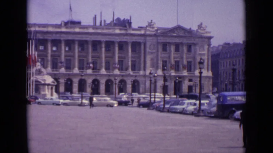1962 Paris frame of a grand classical building across an open square, with cars, pedestrians, flags, and ornate street lamps visible.