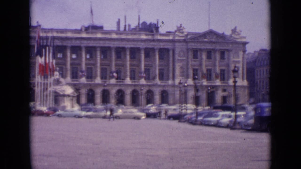 1962 Paris home-movie frame showing a grand stone building, flags, parked cars and pedestrians in a broad square.