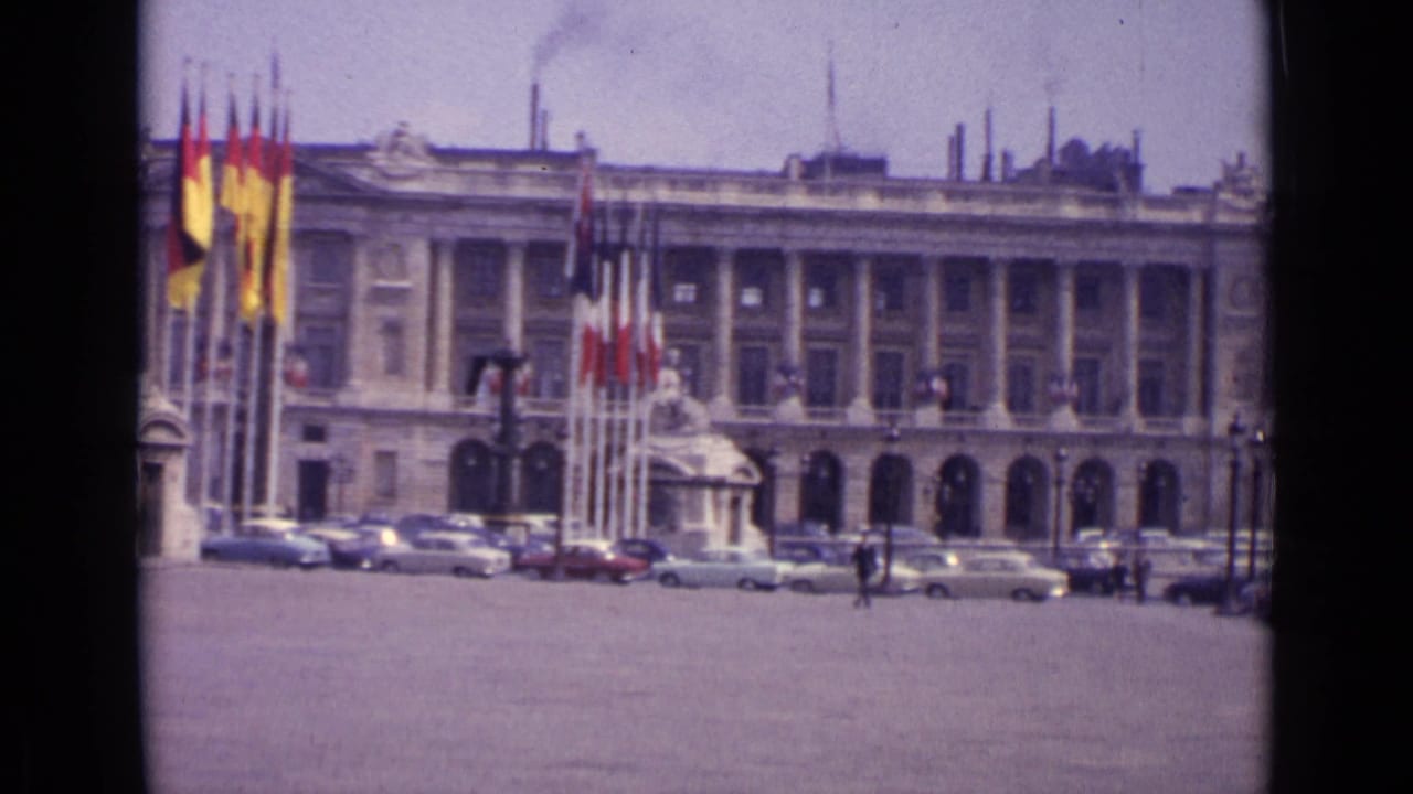 A 1962 Paris view frames flagpoles, parked mid-century cars and a monumental public-looking facade.