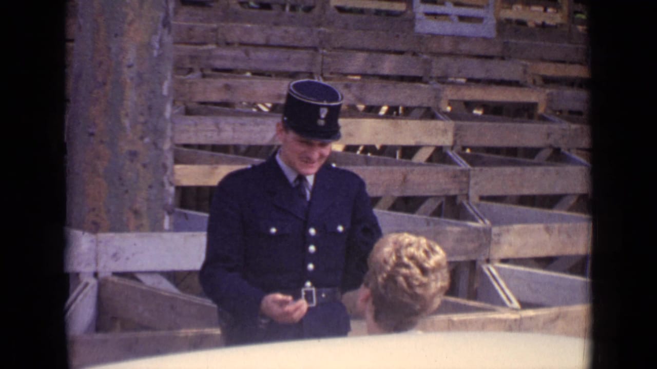 Uniformed policeman faces a blonde woman near stacked wooden crates.