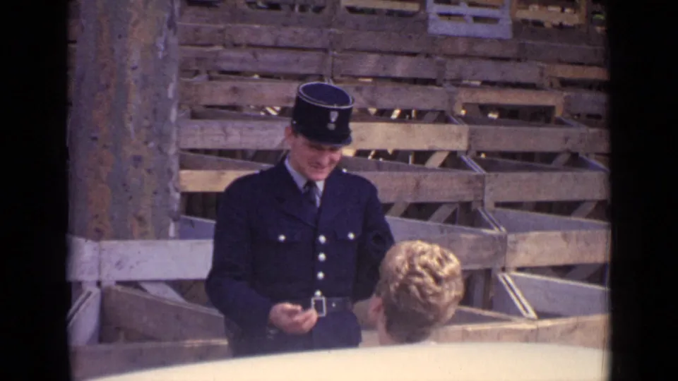 In Paris in 1962, a uniformed policeman in a dark kepi appears to address a blonde woman near stacked wooden crates.