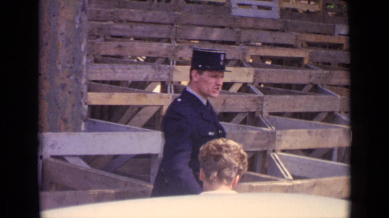 A Paris policeman in dark uniform turns in profile as the woman remains in the foreground near stacked crates.