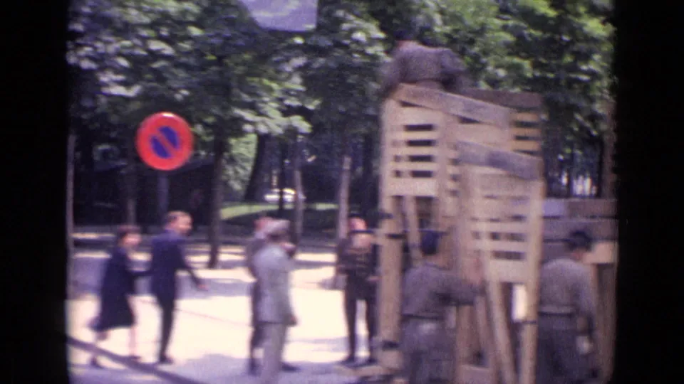 1962 Paris frame with soldiers around a wooden barricade, pedestrians nearby, and a red-and-blue no-parking sign at the curb.