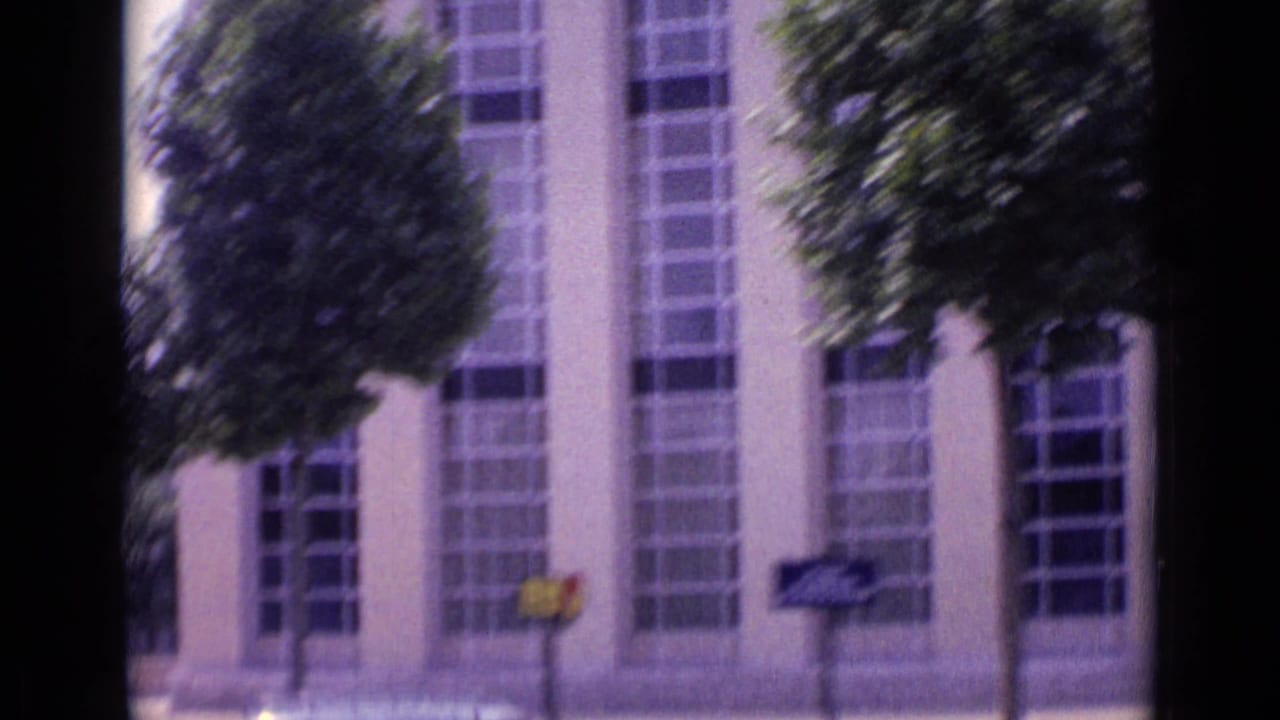 A moving 1962 Paris street view passes tall windows, pale facade panels, trees, and small sidewalk signs.