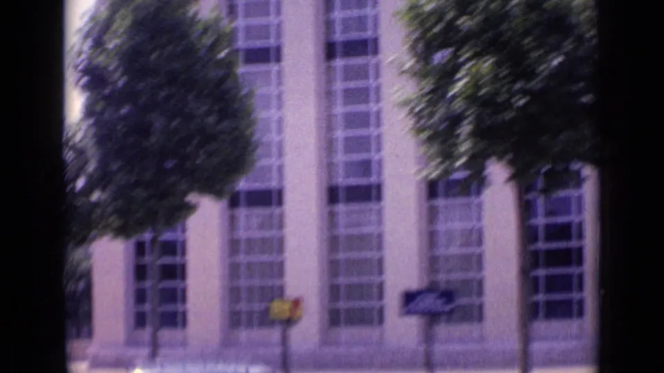 A moving 1962 Paris street view passes tall windows, pale facade panels, trees, and small sidewalk signs.