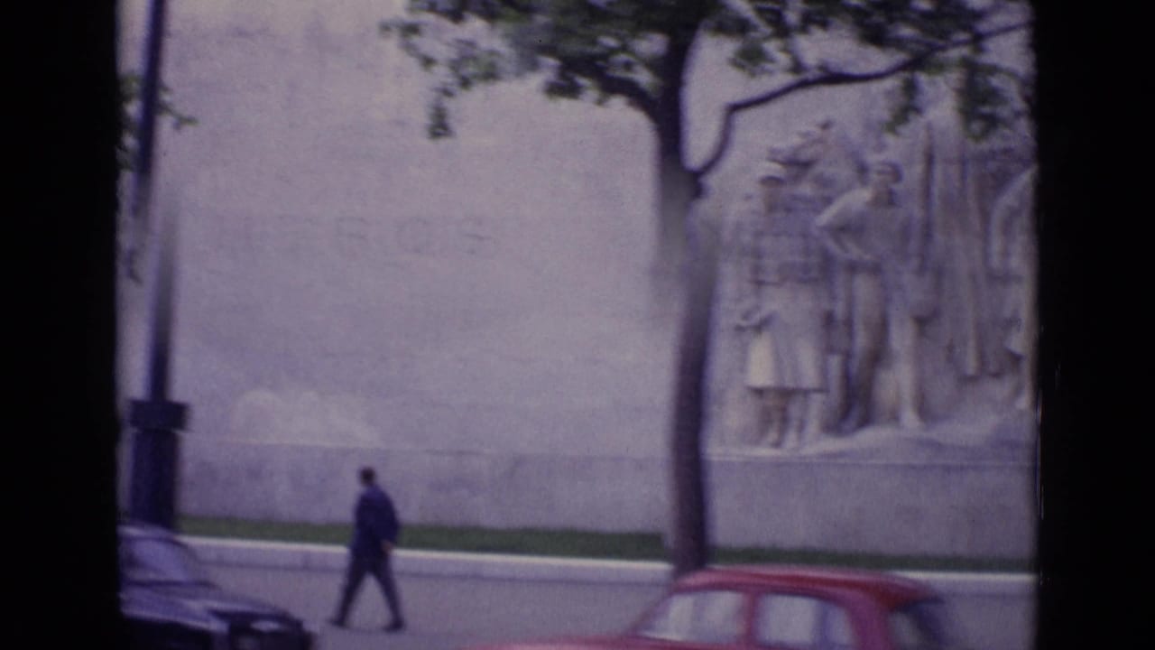 A distant pedestrian walks along a 1962 Paris street beside a pale wall with carved relief figures and parked cars.
